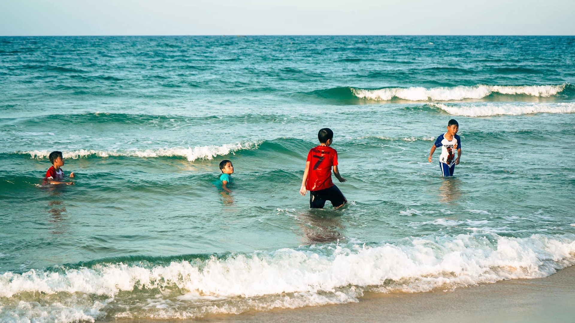 Дети играют с волнами на пляже. Пляж здесь длинный и пологий, морская вода голубая и чистая, безопасная для купания. Дети играют с волнами на пляже. Пляж здесь длинный и пологий, морская вода голубая и чистая, безопасная для купания.